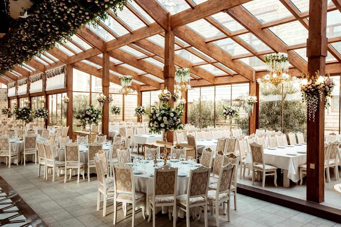 A glass-roofed reception room set with round tables in white linen, white chairs and tall flower arrangements
